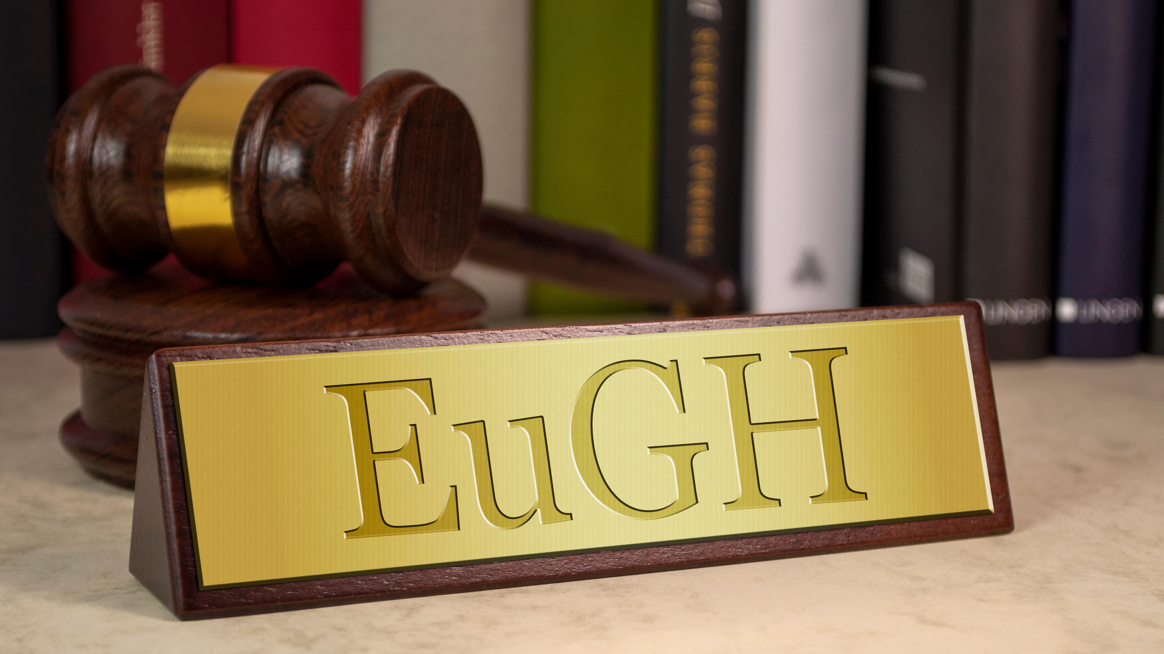 Ein goldenes Schild mit der Aufschrift EuGH steht auf einem Mamor-Tisch. Im Hintergrund sieht man einen Richter-Hammer und einige Bücher.