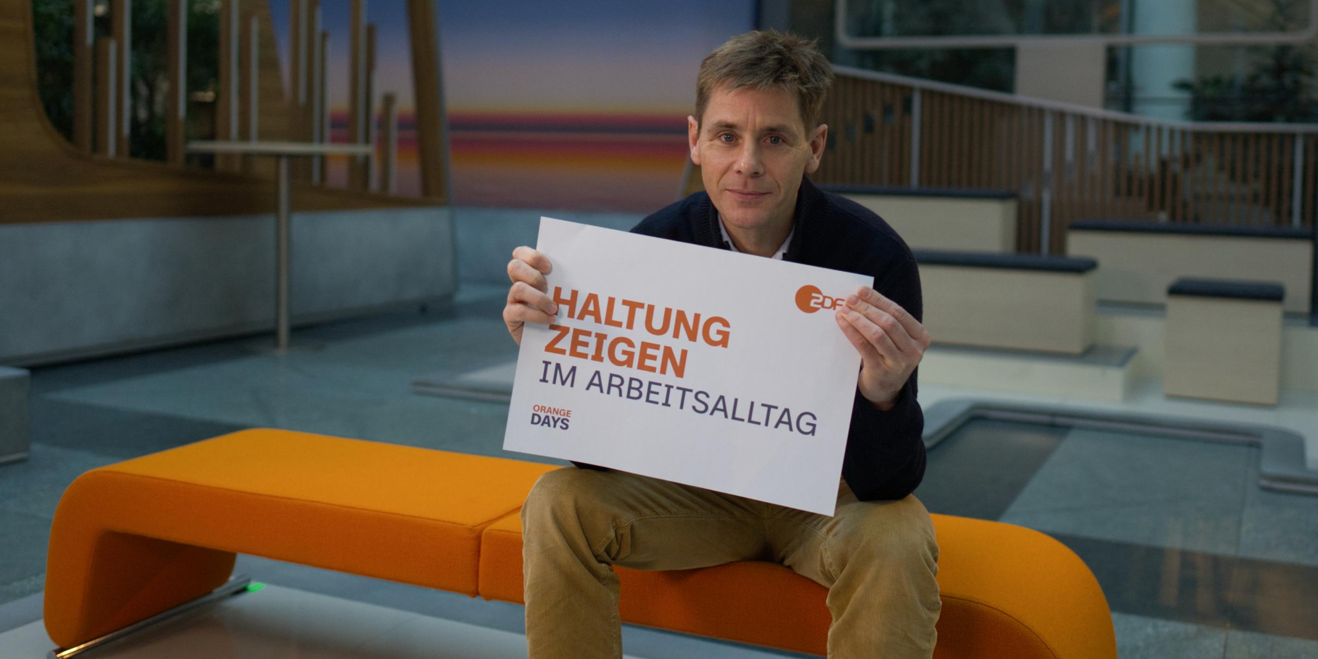 ZDF-Mitarbeiter des ZDF-Studios in Berlin hält Schild zum Aktionstag "Orange Days" mit der Aufschrift "Haltung zeigen im Arbeitsalltag" in die Kamera.