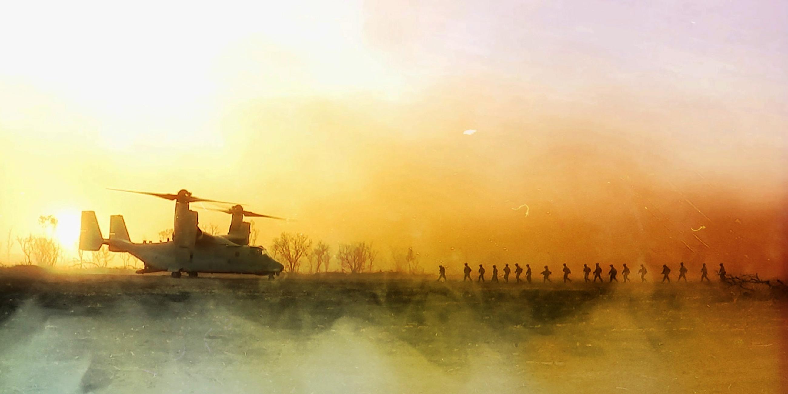 Ein Hubschrauber steht bei Sonnenuntergang auf einem freien Feld. Mehrere Soldaten laufen auf den Hubschrauber zu.