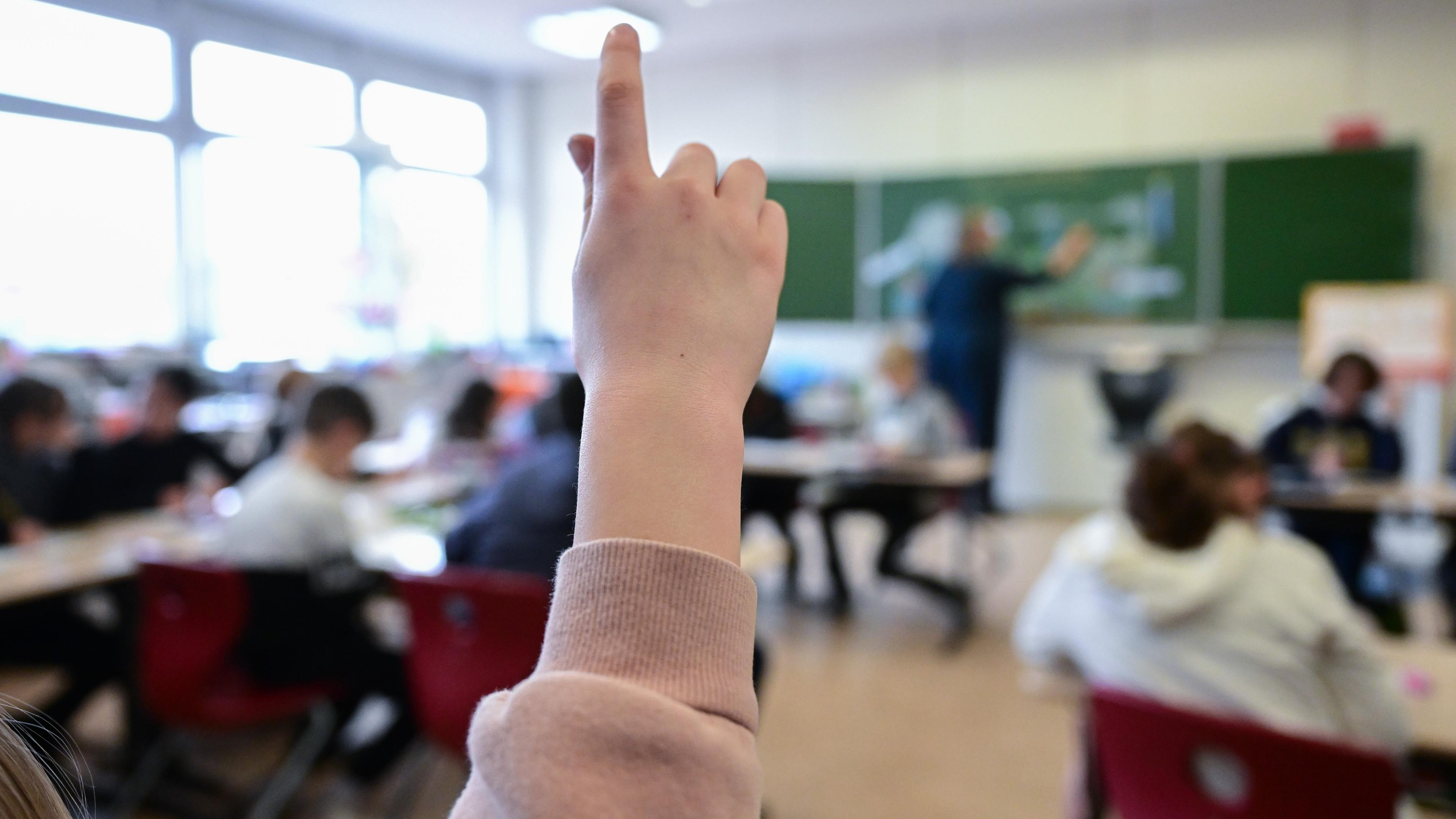 im Vordergrund ist ein Arm von einem Mädchen zu sehen, die sich meldet. Im HIntergund ist verschwommen die restliche Klasse mit Schüler*innen und Lehrerin zu sehen.