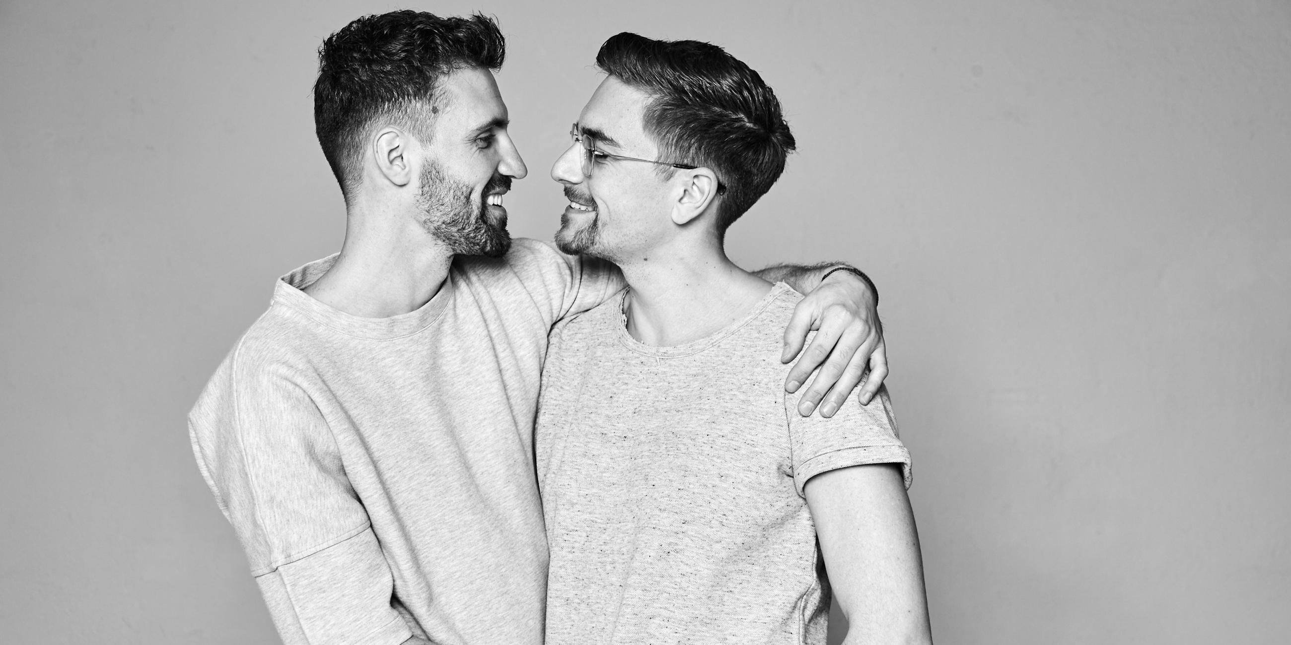 Benjamin und Christophe tragen beide graue T-Shirts und halten sich in den Armen und lächeln sich an. Das Foto ist schwarz-weiß.