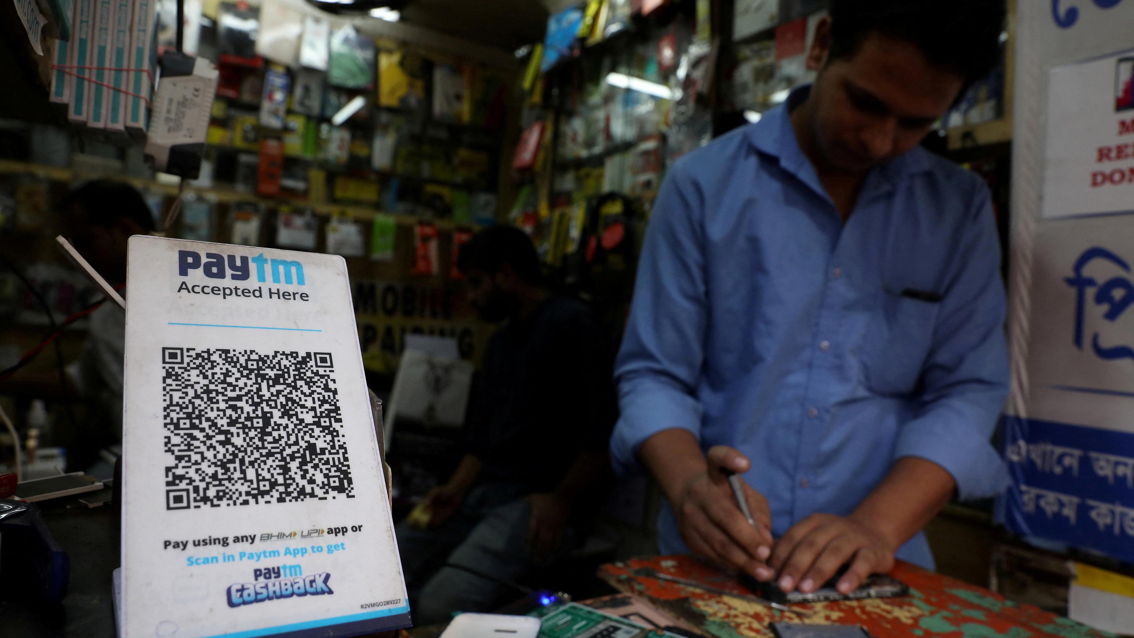 Ein QR-Code ist neben einem Mann abgebildet der hinter einer Kasse steht