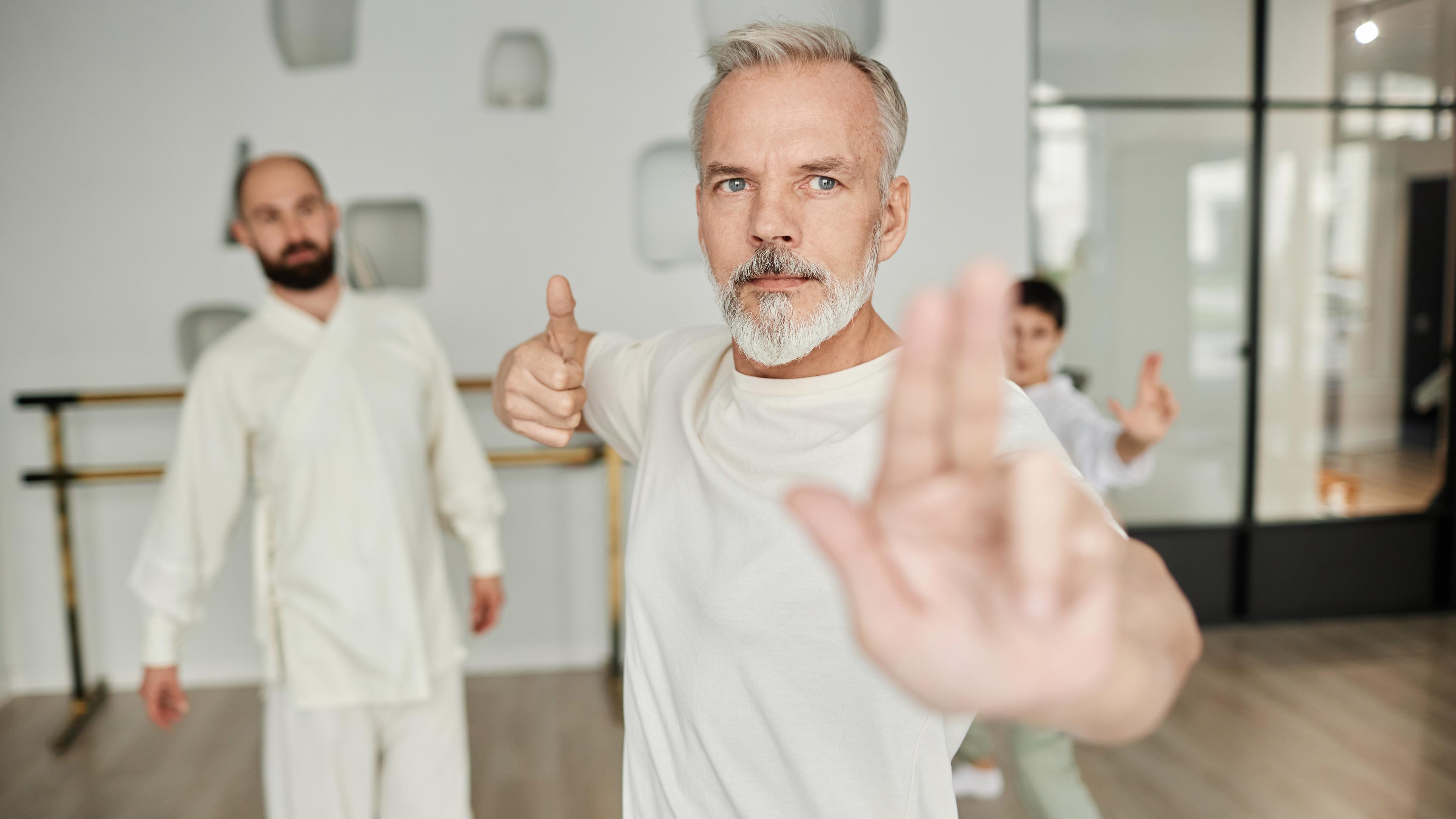 Mann in einer Qigong-Pose, die von einem Trainer im Hintergrund begutachtet wird.