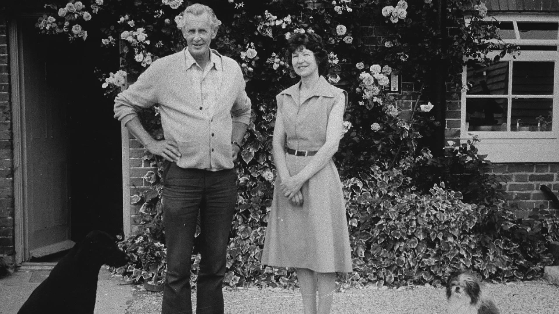 Aus dem Familiendrama "White House Farm": Nevill und June Bamber stehen vor einem mit Kletterrosen bewachsenen Haus und schauen lächelnd in die Kamera. Neben ihnen sitzt ein kleiner Hund.